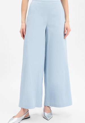 Frau, die hellblaue, weit geschnittene Hosen und silberne, spitze Slingback-Schuhe trägt, steht vor einem einfarbigen Hintergrund.