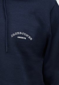 Μπλε σκούρο hoodie με λευκό κείμενο «JACK & JONES ARCHIVE» στο στήθος και ορατό κορδόνι στη αριστερή πλευρά.