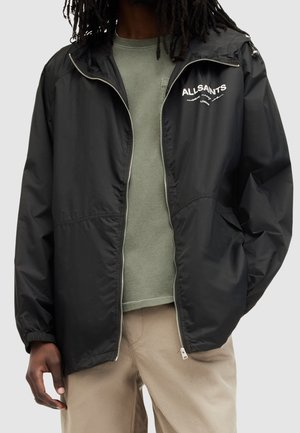 Personne portant une veste zippée noire AllSaints par-dessus une chemise verte et un pantalon beige, debout avec une main dans la poche sur un fond uni.
