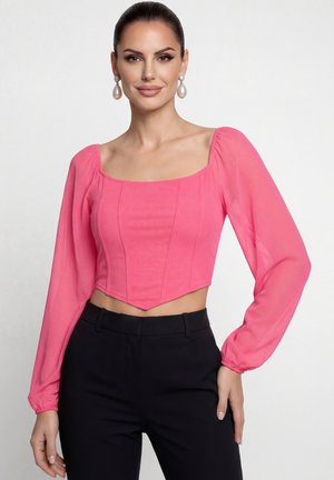 Žena v jasně růžovém krátkém topu s dlouhými rukávy a čtvercovým výstřihem, černých kalhotách s vysokým pasem, s perlovými náušnicemi na zavěšení a se svázanými vlasy.