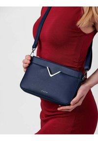 Námořnická modrá syntetická crossbody taška s plochým designem, stříbrným V-článkovým akcentem, nastavitelným popruhem a embosovaným logem na přední straně.