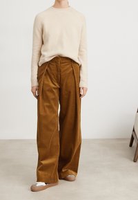 Persona con maglione di maglia beige, pantaloni a gamba larga in corduroy marrone e sneaker beige in piedi su un pavimento chiaro vicino a una sedia di legno.