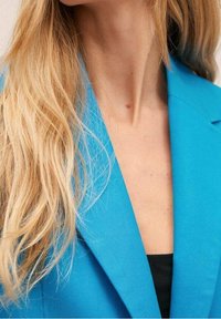 Blazer bleu vif avec une coupe sur mesure, une texture lisse et un col pointu. Porté sur un haut noir, mettant en valeur des cheveux naturellement raides.