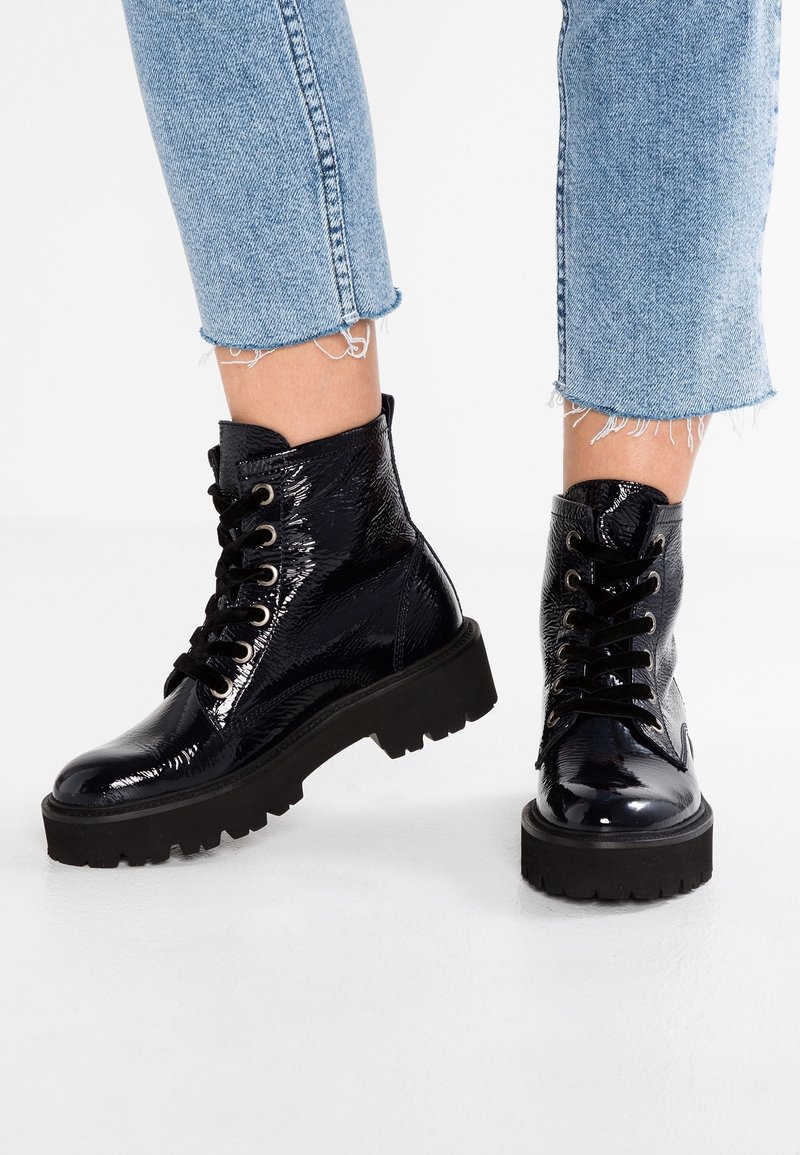 Bottines en cuir verni noir avec une semelle épaisse noire, dotées de lacets ronds et d'une finition texturée. Un jean en denim effiloché est visible au-dessus.