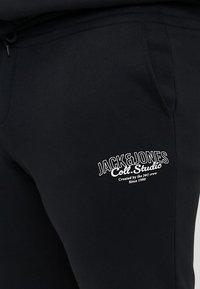 Schwarze Jogginghose aus weichem Material, mit Seitentaschen und einem weißen, gestickten Logo mit Text auf dem linken Oberschenkel.