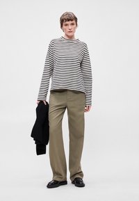 Camisa de manga longa às riscas em preto e branco, combinada com calças largas verde oliva. O modelo segura um suéter de malha preto. Sapatos pretos.