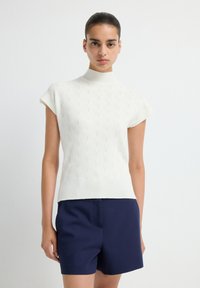 White, short-sleeve knitted turtleneck top with a textured pattern. Worn with dark navy shorts, showcasing a fitted silhouette.