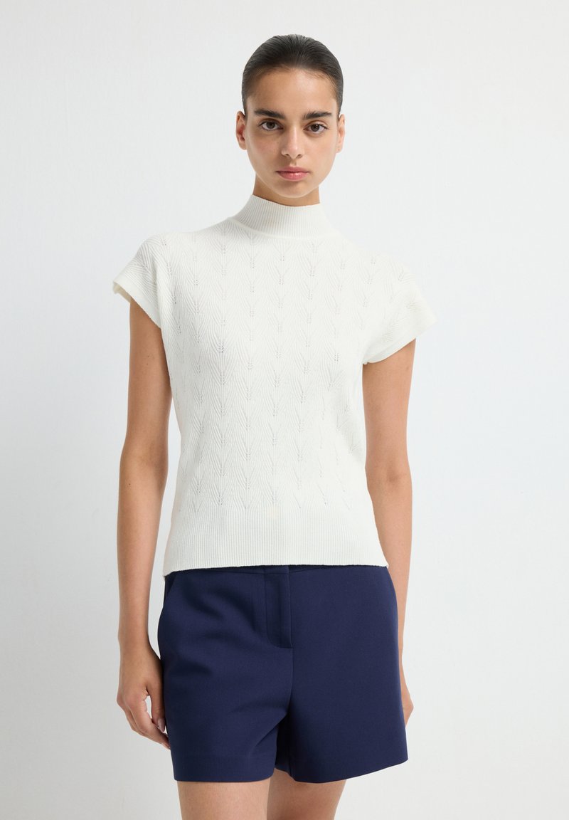 White, short-sleeve knitted turtleneck top with a textured pattern. Worn with dark navy shorts, showcasing a fitted silhouette.