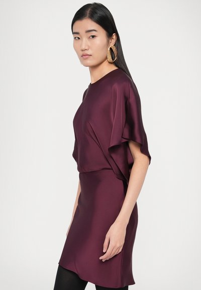 Rochie din satin burgundy cu mâneci scurte și vaporoase și siluetă ajustată. Modelul poartă o cercească mare aurie, stilizată cu ciorapi negri.