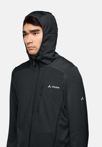 Giacca nera con cappuccio e finitura testurizzata, dotata di zip frontale, tasche laterali e logo Vaude sul petto e sulla manica.