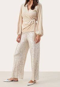 Top envolvente beige con mangas largas abullonadas, combinado con pantalones de pierna ancha cubiertos de lentejuelas plateadas. Zapatos planos plateados reflectantes completan el look.