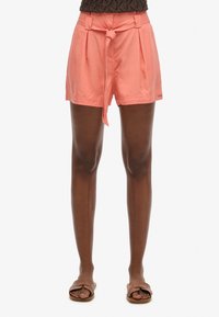 Peach-colored shorts with pleated front and fabric tie at the waist. Smooth texture, knee-length, paired with brown flat sandals.