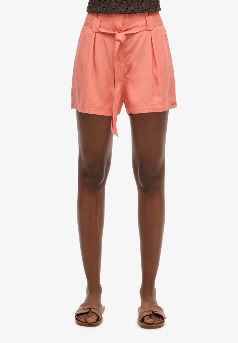 Peach-colored shorts with pleated front and fabric tie at the waist. Smooth texture, knee-length, paired with brown flat sandals.
