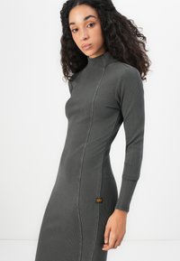 Šaty s dlhým rukávom v tmavošedej rebrovanej tkanine, s vysokým golierom, priliehavým strihom a jemnou značkovou etiketou na boku.