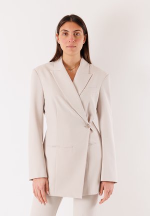 Donna con i capelli lunghi e scuri indossa un blazer beige oversize con un solo bottone, in piedi davanti a uno sfondo bianco uniforme.