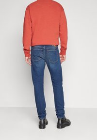 Sudadera roja, vaqueros azules y botas negras. Los vaqueros tienen bolsillos traseros y un diseño de canesú curvado, con detalles de costura visibles.