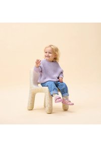 Suéter de lana lila, jeans de mezclilla y zapatos rosas en un niño sentado en una silla de plástico moteada. Fondo beige claro.