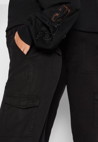 Pantalons cargo noirs avec poches latérales et un tissu texturé, associés à un haut noir à manches longues avec une broderie sur la manche.