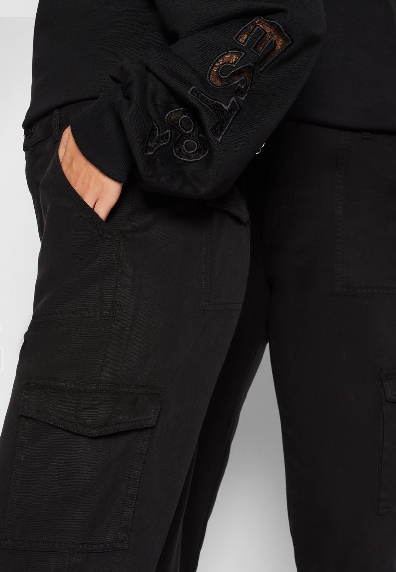 Pantalons cargo noirs avec poches latérales et un tissu texturé, associés à un haut noir à manches longues avec une broderie sur la manche.