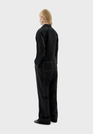 Jumpsuit di denim nero con maniche lunghe, tasche laterali e cuciture gialle a contrasto, indossato con scarpe nere. Vista da dietro.