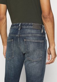 Dunkelblaue Jeans aus Denim mit schmaler Passform, ausgestattet mit Gesäßtaschen, kontrastierendem Nähen und einem gebrandeten Lederpatch am Bund.