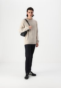 Bež oversized pulover s teksturiranim pletenjem, crna torba preko ramena, tamne hlače i crne tenisice s bijelim prugama. Jednostavan, moderan dizajn.