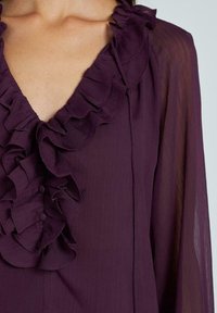 Blusa viola con scollatura a V profonda e rifiniture a volant, caratterizzata da un tessuto morbido e bottoni decorativi disposti lungo la parte frontale.