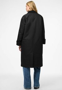 Lungo cappotto nero con colletto, spacco posteriore e maniche ampie, abbinato a jeans blu a gamba larga. Tessuto morbido, design minimale.