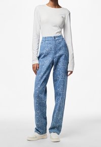 Person i vit långärmad topp, högmidjade blå byxor med rosor, och vita sneakers, stående mot en vit bakgrund.