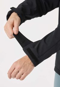 Rankos reguliuoja juodo švarko rankovės užsegimą su „Velcro“ dirželiu prieš balto paprasto fono foną.