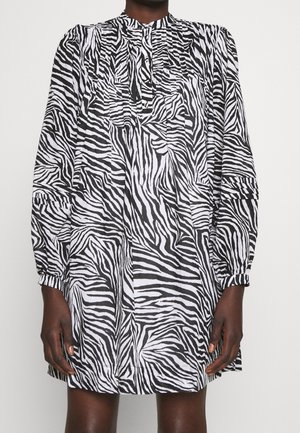 Person iført en sort og hvid zebra-print langærmet kjole med rynkede ærmer og høj halsudskæring mod ensfarvet baggrund.
