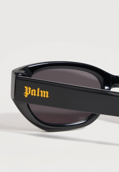 Lunettes de soleil noires avec des montures épaisses et des verres foncés, avec le mot « Palm » en police gothique jaune sur la branche.