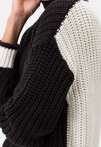 Sweter z dzianiny o czarnej korpusie i białych rękawach, z kontrastującymi ściągaczami. Teksturalna tkanina o luźnym kroju i okrągłym dekolcie.