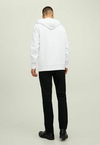 Weißer Kapuzenpullover mit langen Ärmeln, ausgestattet mit einer Kängurutasche und gerippten Bündchen. Kombiniert mit schwarzen Slim-Fit-Hosen und schwarzen Stiefeln.