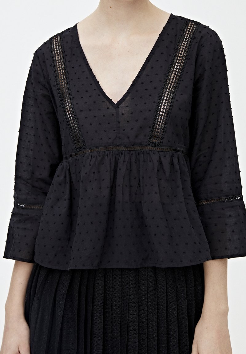 Blusa nera, testurizzata, con scollatura a V e maniche a tre quarti. Presenta un bordo in pizzo e una vita arricciata. Realizzata in tessuto leggero.