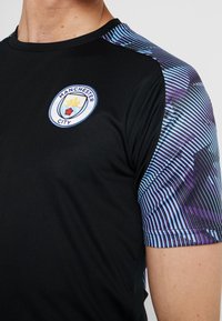 Mustapaita, jossa on pyöreä pääntie. Hihassa sinivioletteja raitoja. Rinnassa on ympyränmuotoinen Manchester City -logo.