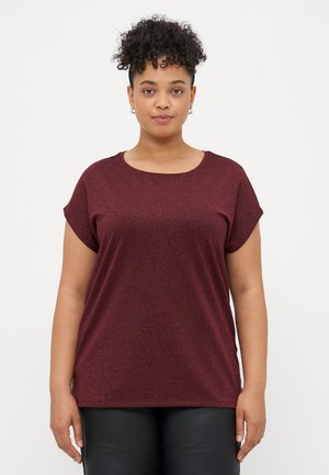 Vrouw met krullend haar, gekleed in een kort mouwige bordeauxrode glinsterende top en zwarte broek, staand tegen een effen witte achtergrond.