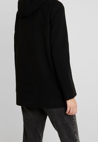 Abrigo negro con capucha, de diseño recto y mangas largas, con tejido texturizado. Lleva puesto sobre unos vaqueros oscuros de pierna recta.