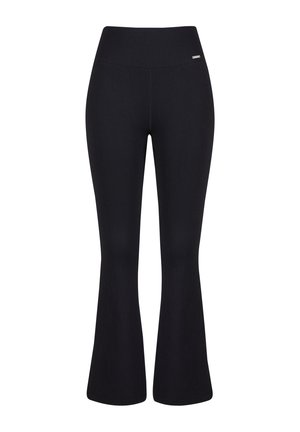 Femme portant un pantalon évasé côtelé noir taille haute, un crop top assorti sans manches et des baskets blanches, debout devant un fond blanc.
