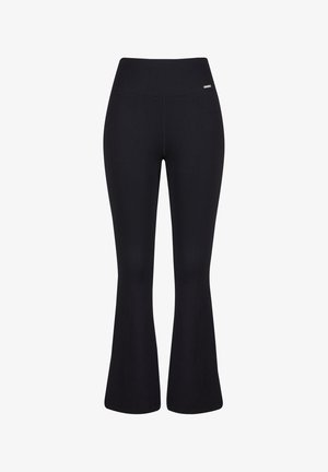 Femme portant un pantalon évasé côtelé noir taille haute, un crop top assorti sans manches et des baskets blanches, debout devant un fond blanc.