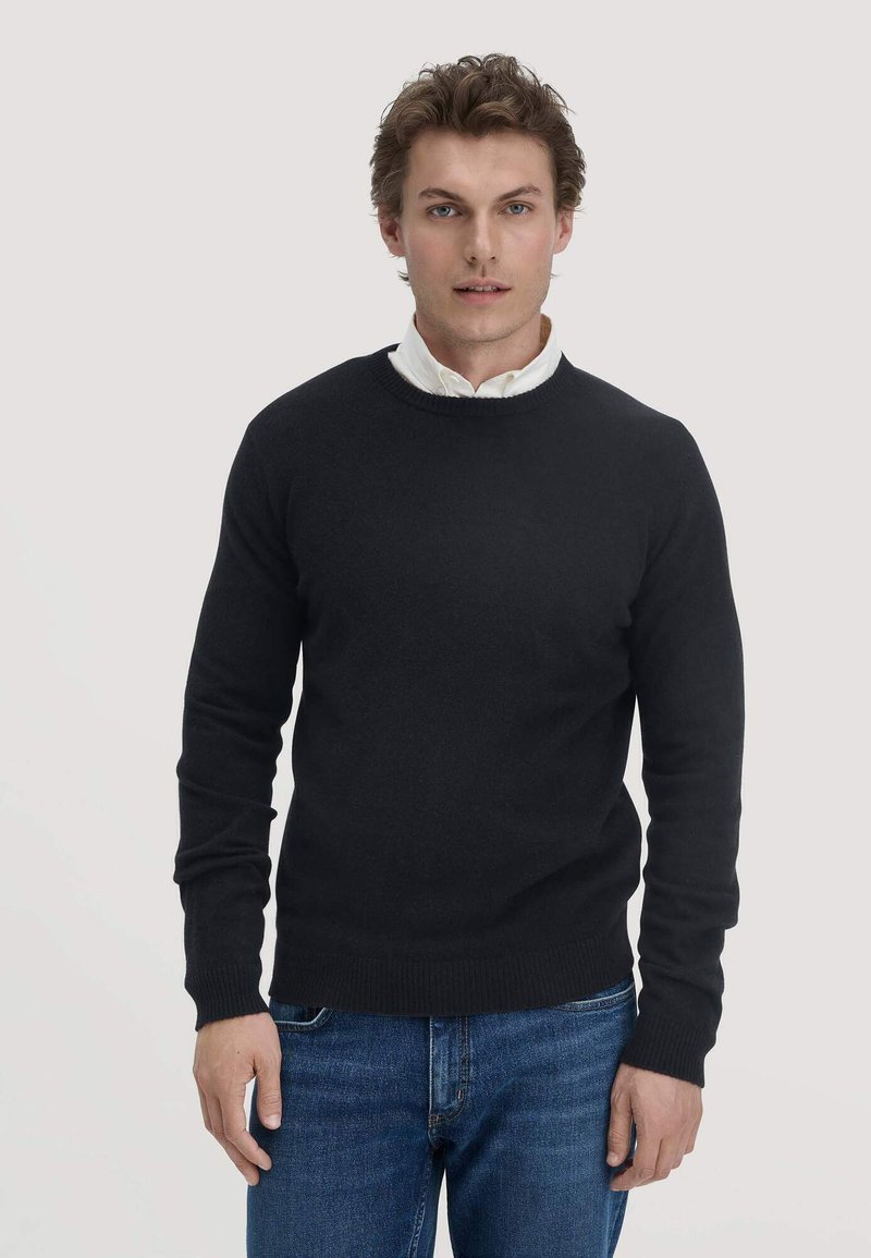 Schwarzer Strickpullover mit Rundhalsausschnitt und gerippten Bündchen, über einem weißen Hemd mit Kragen getragen. Slim Fit mit glatter Textur. Jeans sichtbar.