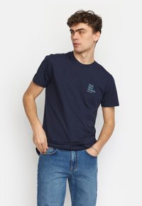 Marineblauwe katoenen T-shirt met korte mouwen en een ronde hals, voorzien van teal borduurwerk van "Fresh Meat Mondays" op de linkerkant van de borst.