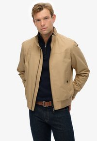 Lichte tan jack met een staande kraag, ritssluiting en zijzakken. Draagt over een marineblauwe pullover en is gecombineerd met donkere jeans.