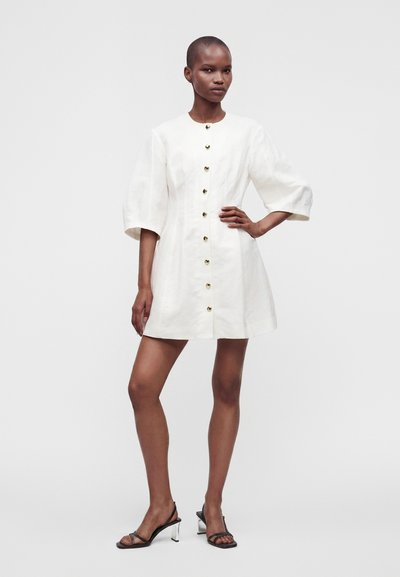 Femme portant une robe blanche boutonnée avec des manches bouffantes trois-quarts et des sandales à talons noires à lanières, debout avec une main sur la hanche.