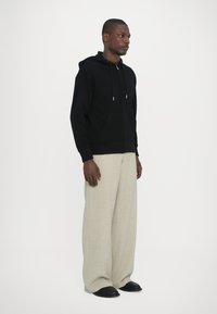Felpa nera con cappuccio e lacci, abbinata a pantaloni ampi beige chiaro e scarpe nere. Tessuto morbido al tatto, vestibilità casual.