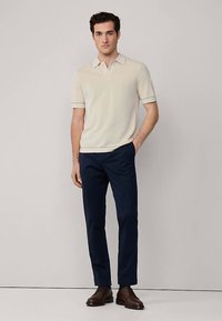 Beige värinen lyhythihainen poolopaita, jossa on kaulus. Yhdistetty tummansinisiin housuihin ja tummanruskeisiin kenkiin. Sileä pinta ja vartalonmyötäinen malli.