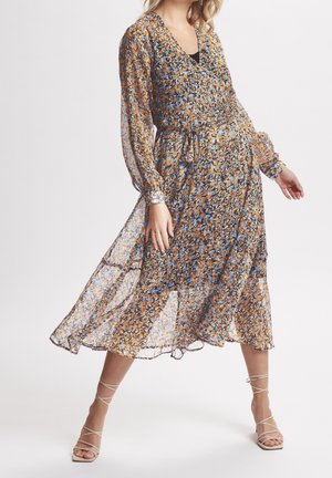 Vrouw draagt een vloeiende midi-jurk met lange mouwen in oranje, blauw en zwart bloemenpatroon, gecombineerd met beige sandalen met hak en bandjes.