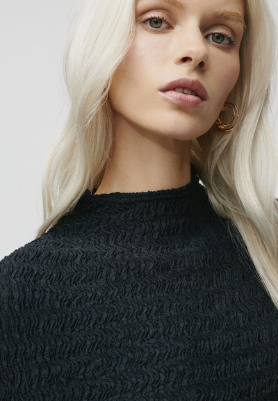 Top nero in tessuto testurizzato con un design ondulato e scollatura alta. Il modello ha lunghi capelli biondi lisci e orecchini a cerchio dorati.