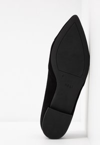 Zapato de tacón de ante negro con punta afilada y suela plana. La suela tiene textura, con una ligera curvatura y el nombre de la marca grabado.