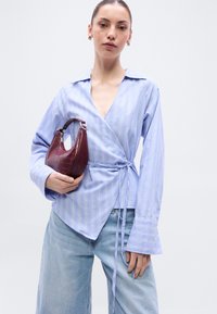 Γαλάζιο πουκάμισο με ρίγες λευκές, μακριά μανίκια και ζωνάκια στο λαιμό. Το μοντέλο κρατάει μια μπορντό τσάντα με σχέδιο κροκοδείλου. Φοράει γαλάζια τζιν παντελόνια.